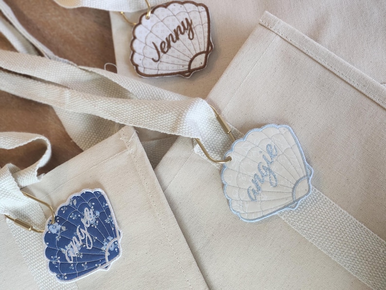 May include: Close-up of three canvas tote bags with seashell-shaped name tags. The tags are in various colors, including blue and cream, and feature names like "Jenny" and "Angie" in cursive script. The bags have cream-colored straps.