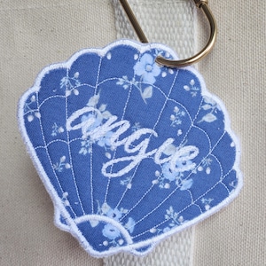 May include: A blue seashell-shaped embroidered patch with the name "Angie" in white script. The patch features a floral pattern and a white scalloped border. Attached to a gold carabiner clip and a cream-colored tote bag.