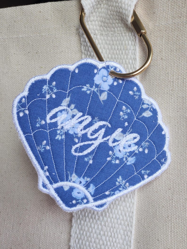 May include: A blue seashell-shaped embroidered patch with the name "Angie" in white script. The patch features a floral pattern and a white scalloped border. Attached to a gold carabiner clip and a cream-colored tote bag.