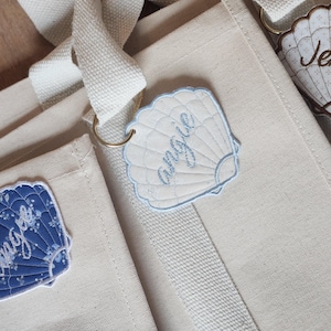 May include: Close-up of three canvas tote bags with seashell-shaped name tags. The tags are white, blue, and floral patterned, with names embroidered on them. The bags have cream-colored straps and are on a wooden surface.
