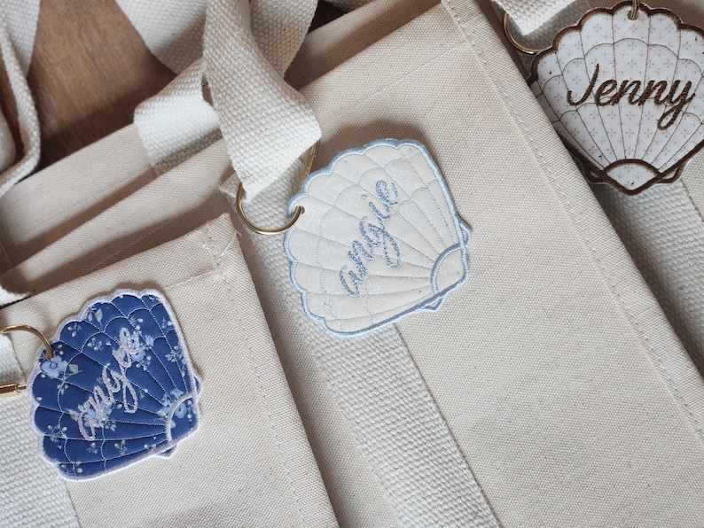 May include: Close-up of three canvas tote bags with seashell-shaped name tags. The tags are white, blue, and floral patterned, with names embroidered on them. The bags have cream-colored straps and are on a wooden surface.