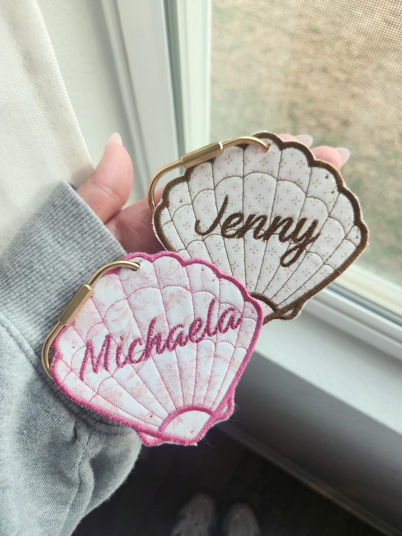 May include: Two seashell-shaped luggage tags. One is white with brown trim and the name "Jenny" embroidered in brown. The other is pink with the name "Michaela" embroidered in pink. Both have gold-colored metal clasps.