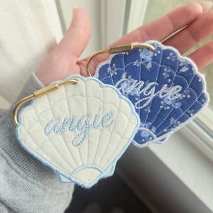 May include: Two seashell-shaped keychains with the name "Angie" embroidered in light blue. One keychain is white with a light blue border, and the other is blue with a floral pattern. Both have gold-colored metal clasps.