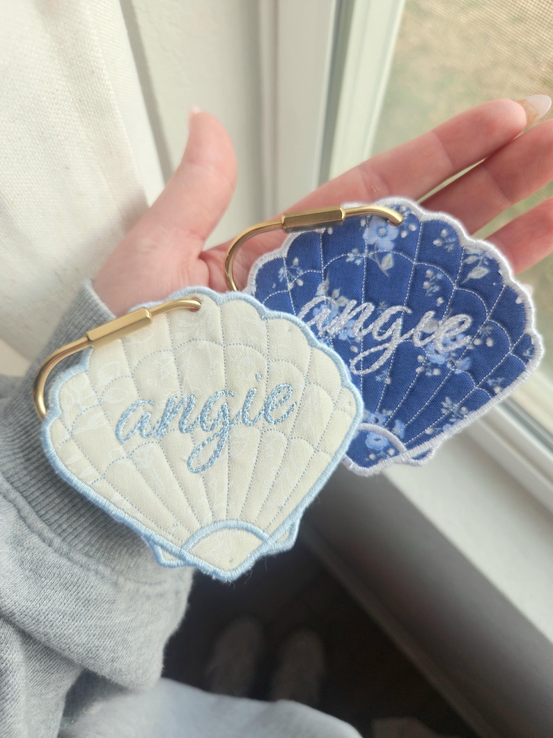 May include: Two seashell-shaped keychains with the name "Angie" embroidered in light blue. One keychain is white with a light blue border, and the other is blue with a floral pattern. Both have gold-colored metal clasps.