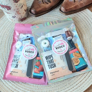 May include: Two Last Rodeo Hangover Kits in pink and iridescent pouches, each containing wipes, mints, and other essentials. The kits are displayed with cowboy boots on a woven mat, suggesting a rodeo theme.
