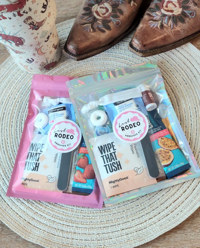 May include: Two Last Rodeo Hangover Kits in pink and iridescent pouches, each containing wipes, mints, and other essentials. The kits are displayed with cowboy boots on a woven mat, suggesting a rodeo theme.