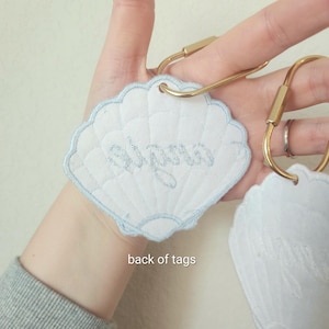 May include: White seashell-shaped tags with light blue embroidered trim and text. The tags are attached to gold-colored key rings. The text "Alyssa" is embroidered on one tag. The back of the tags is visible in the image.