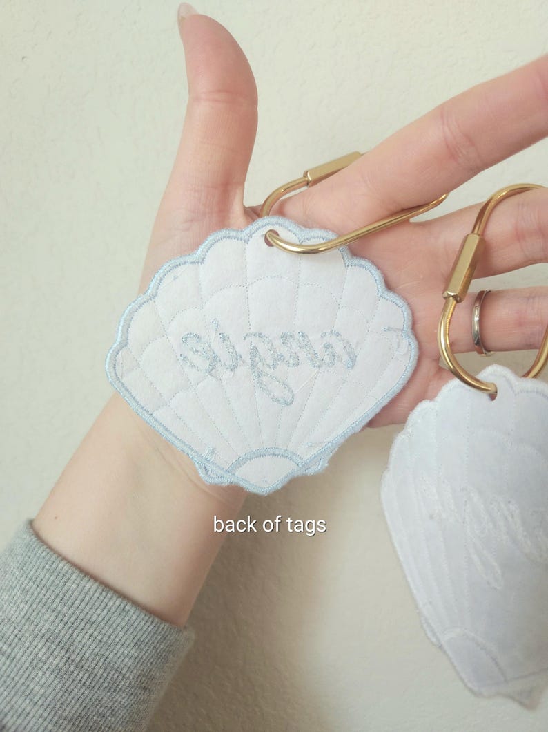 May include: White seashell-shaped tags with light blue embroidered trim and text. The tags are attached to gold-colored key rings. The text "Alyssa" is embroidered on one tag. The back of the tags is visible in the image.