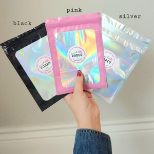 May include: Three colorful, holographic pouches are held up against a light background. The pouches are labeled "black", "pink", and "silver". Each pouch has a circular sticker with the text "Lash Rodeo Hangover Kit". The pink pouch is held by a person wearing a denim jacket.