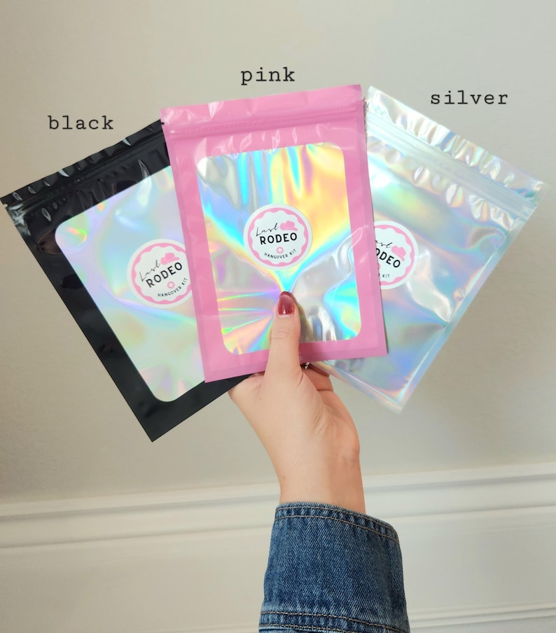 May include: Three colorful, holographic pouches are held up against a light background. The pouches are labeled "black", "pink", and "silver". Each pouch has a circular sticker with the text "Lash Rodeo Hangover Kit". The pink pouch is held by a person wearing a denim jacket.