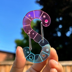 以下が含まれることがあります： 「S」の形をした、カラフルな虹色の透明アクリルチャーム。ピンクからブルーに色が変化し、星と月の切り抜きがあります。シルバーカラーの留め金が付いています。