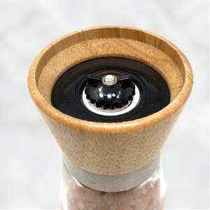 Peut inclure: Gros plan sur un moulin &agrave; sel en bois avec un m&eacute;canisme de broyage noir. Le dessus est en bois clair et le corps en verre transparent rempli de sel rose. Une bande argent&eacute;e entoure le verre et le bois.