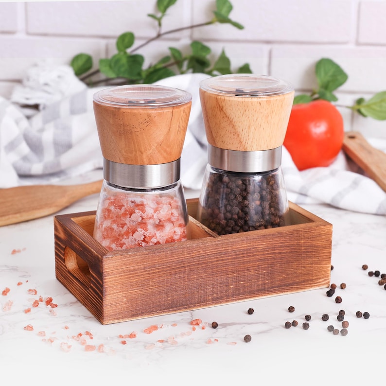 Peut inclure: Un ensemble de moulins &agrave; sel et &agrave; poivre en bois. Les moulins ont des bases en verre, des bandes en acier inoxydable et des dessus en bois. L'un contient du sel rose de l'Himalaya, l'autre des grains de poivre noir. L'ensemble est dans un plateau en bois teint&eacute;.