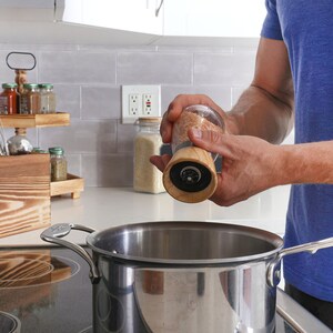 Peut inclure: Un moulin &agrave; sel en verre et bois est tenu au-dessus d'une casserole en acier inoxydable sur une cuisini&egrave;re. Le moulin contient du gros sel. Un pr&eacute;sentoir &agrave; &eacute;pices et un porte-ustensiles en bois sont visibles en arri&egrave;re-plan, dans une cuisine.