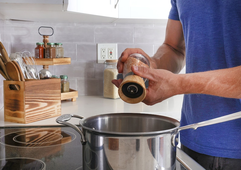Peut inclure: Un moulin &agrave; sel en verre et bois est tenu au-dessus d'une casserole en acier inoxydable sur une cuisini&egrave;re. Le moulin contient du gros sel. Un pr&eacute;sentoir &agrave; &eacute;pices et un porte-ustensiles en bois sont visibles en arri&egrave;re-plan, dans une cuisine.