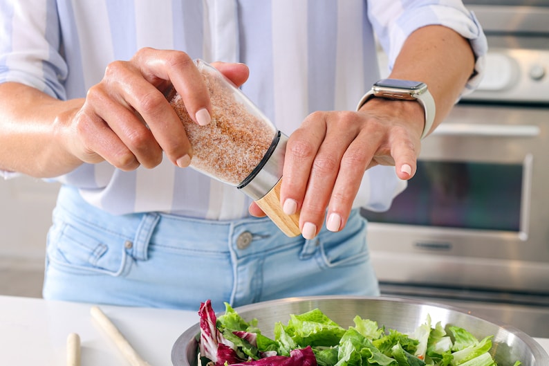 Peut inclure: Un sali&egrave;re en verre transparent avec une base en bois et un bord en acier inoxydable est utilis&eacute; pour assaisonner une salade. La sali&egrave;re contient du sel brun clair grossier. La salade est dans un bol en acier inoxydable avec diverses verdure.