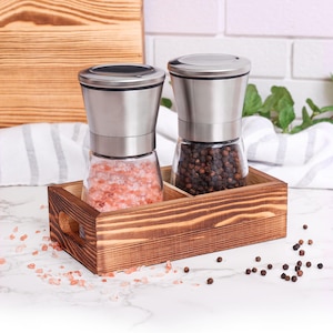 May include: Two stainless steel salt and pepper grinders with glass containers filled with pink Himalayan salt and black peppercorns. The grinders are sitting in a wooden tray.