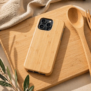 Puede incluir: Una funda de teléfono de bambú marrón claro con una textura de vetas de madera natural. La funda está sobre una tabla de cortar de madera con una cuchara y un tenedor de madera. La funda del teléfono tiene un módulo de cámara negro.