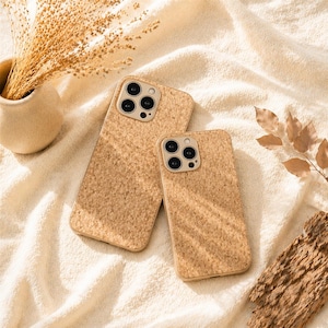 Könnte beinhalten: Zwei beige Kork-Handyhüllen mit Kameraausschnitten liegen auf einer weißen, strukturierten Oberfläche. Die Hüllen haben ein natürliches, strukturiertes Aussehen. Ein kleiner Vasen mit getrockneten Pflanzen und ein Stück Rinde sind ebenfalls im Bild.