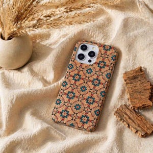 Può includere: Custodia per telefono con motivo floreale nei toni del verde acqua, rosso e marrone. La custodia è realizzata in un materiale naturale e si trova su un tessuto beige. Sono visibili anche un piccolo vaso con erba secca e pezzi di corteccia.