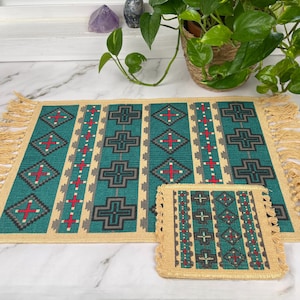 Op de afbeelding: Een geweven placemat- en onderzetterset. Het turquoise, beige, rode en grijze ontwerp heeft geometrische patronen en franjes. De set wordt gepresenteerd op een wit marmeren oppervlak, met een kamerplant op de achtergrond.