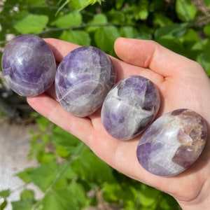May include: Four smooth, polished amethyst tumbled stones in various shades of purple and white. The stones are arranged in a row on a hand.