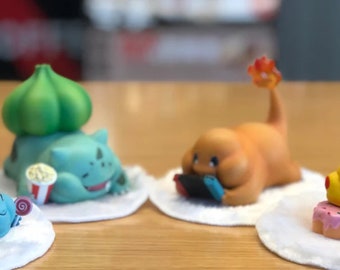 May include: Four Pokemon figurines on white bases. A green and blue Squirtle figurine is eating a donut. A brown and blue Squirtle figurine is eating a donut. A yellow Pikachu figurine is eating a donut. A brown and orange Charmander figurine is holding a video game controller.