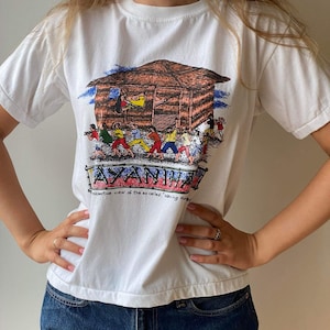 Può includere: Maglietta bianca con un'illustrazione colorata di una capanna e persone. Il testo "AYANHIA" e "Perspective view of the so called 'haring house'" è stampato sulla maglietta.