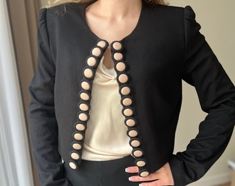 May include: A black cropped blazer with a round neckline and long sleeves. The blazer has a row of large, beige buttons down the front.