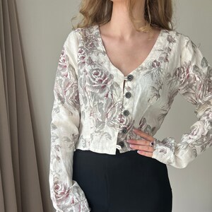 Könnte beinhalten: Eine weiße Leinenbluse mit einem Blumendruck mit grauen und burgunderfarbenen Rosen. Die Bluse hat einen V-Ausschnitt und eine Knopfleiste vorne.