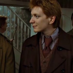 Puede incluir: Un hombre con cabello castaño rojizo, vistiendo un abrigo marrón, una camisa estampada y una corbata burdeos. Mira hacia un lado con una leve sonrisa. Otro hombre es visible en el fondo.