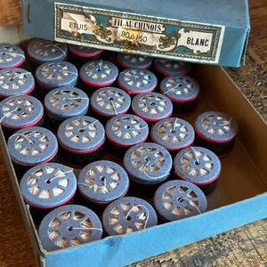 May include: A blue box filled with spools of white thread. The box has a label that reads "FIL AU CHINOIS 80 a 150 BLANC".