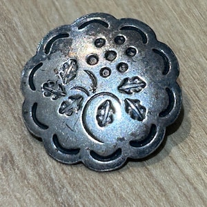May include: A silver-toned metal button with a scalloped edge and a floral design. The button features leaves, a crescent moon, and small circular details. The button is circular and has a slightly aged appearance.