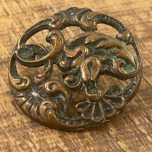 May include: A circular metal button featuring an openwork design with intricate, raised scrollwork and floral motifs. The surface has a weathered, dark bronze finish with visible green oxidation in the recessed areas of the metalwork.