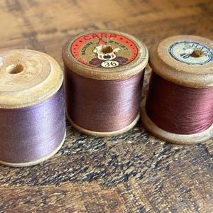 May include: Three vintage wooden thread spools in shades of purple and brown. The spools have paper labels with text and numbers. The thread is tightly wound around the wooden cores, showcasing the colours and textures of the thread.