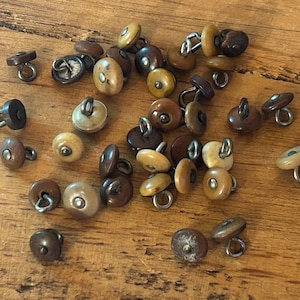 Könnte beinhalten: Eine Sammlung von Vintage-Knöpfen in verschiedenen Braun- und Cremetönen mit Metallstiften. Die Knöpfe sind auf einer Holzoberfläche verstreut und zeigen ihre unterschiedlichen Formen und Größen. Diese Knöpfe eignen sich ideal für Näh- und Bastelprojekte.