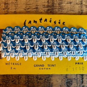 May include: A yellow card with the word "Fantasie" printed in black. The card has a blue and white trim with a pattern of loops. The card also has the text "Mètrage 2 m. Grand Teint Coton Prix #1F50".
