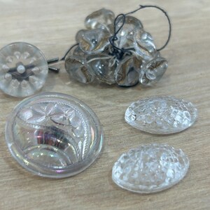 May include: A collection of clear glass buttons featuring various textures and patterns. One button has a floral design, two are oval with a textured surface, and several others are round with a sunburst pattern, some attached with black thread.