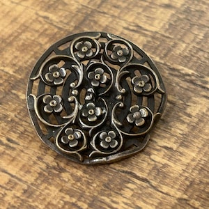 May include: A circular metal button featuring an intricate openwork design of swirling vines and small four-petaled flowers. The metal has a dark, aged finish with polished highlights on the floral elements and a central shank.