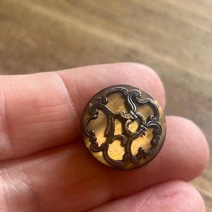 May include: A round button featuring a dark brown, translucent base with an intricate, raised metallic overlay in a swirling, ornate pattern. The metallic design covers the surface of the button, creating a contrast against the smooth, glossy brown material underneath.