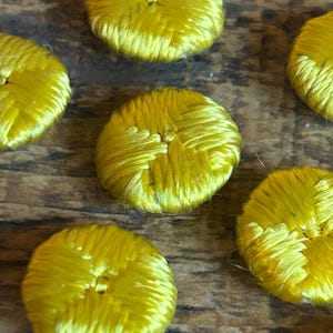 May include: Close-up of several bright yellow, round, fabric-covered buttons. The buttons have a textured, woven appearance and are arranged on a wooden surface. The buttons are a vibrant, sunny yellow colour.