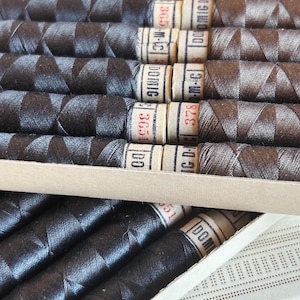 May include: Close-up of a box of spools of thread. The spools are wrapped in black and brown thread. The labels on the spools read "DOMIG 3651", "GD-M 3780-M-C", "DOMIG 365", and "IC-D-M 3865".