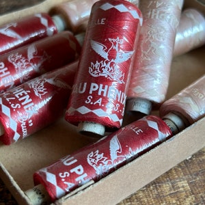 May include: A box filled with spools of red and pink thread. The spools are labeled "AU PHENIX S.A."