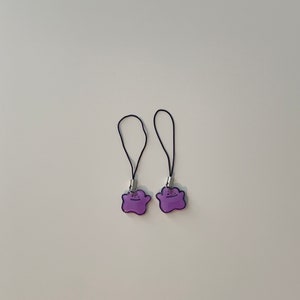 May include: Two purple and black phone charms with a cartoon ghost design. The charms are attached to black strings.