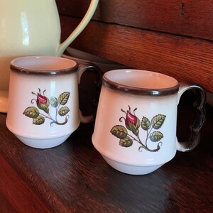 May include: Two white ceramic mugs with brown rims and handles. Each mug features a floral design with a red rose, green leaves, and brown stems. The mugs are sitting on a wooden surface next to a yellow teapot.