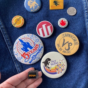 May include: A collection of vintage buttons featuring various designs, including a lion, a castle, a zodiac sign, and a sea world logo. The buttons are displayed on a blue denim background.