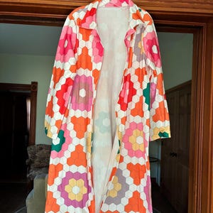 May include: A long, open-front robe with a vibrant geometric pattern in shades of orange, pink, white, and green. The robe is hanging on a hanger, showcasing its full length and design. The pattern is composed of hexagonal shapes.
