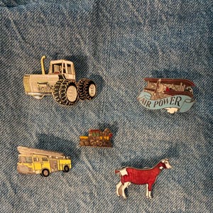 May include: Collection of enamel pins featuring a white tractor, a biplane with "AIR POWER" text, a yellow fire truck, a small train, and a red and white goat. The pins are set against a blue denim background.