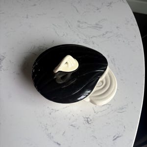 May include: A black and white ceramic container with a lid. The lid is black with a white handle. The container has a circular shape. The container is on a white surface.