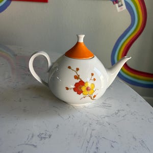 May include: A white teapot with an orange lid and floral design. The teapot has a delicate floral pattern with yellow and orange flowers on a white background.